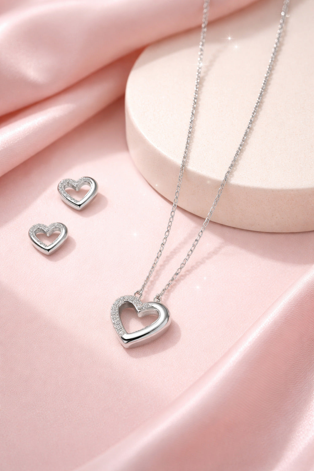 Silver heart-shaped necklace and earrings on a pink fabric background