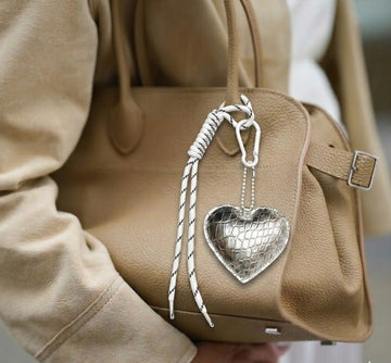 Beige handbag with a silver heart-shaped charm held by a person wearing a beige coat.