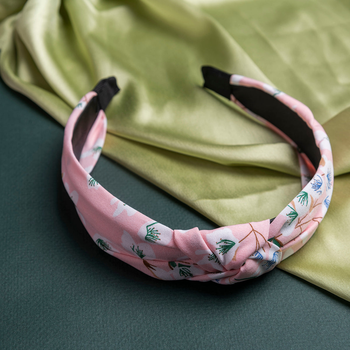 A baby pink hair band with a floral pattern, resting on a green fabric surface.