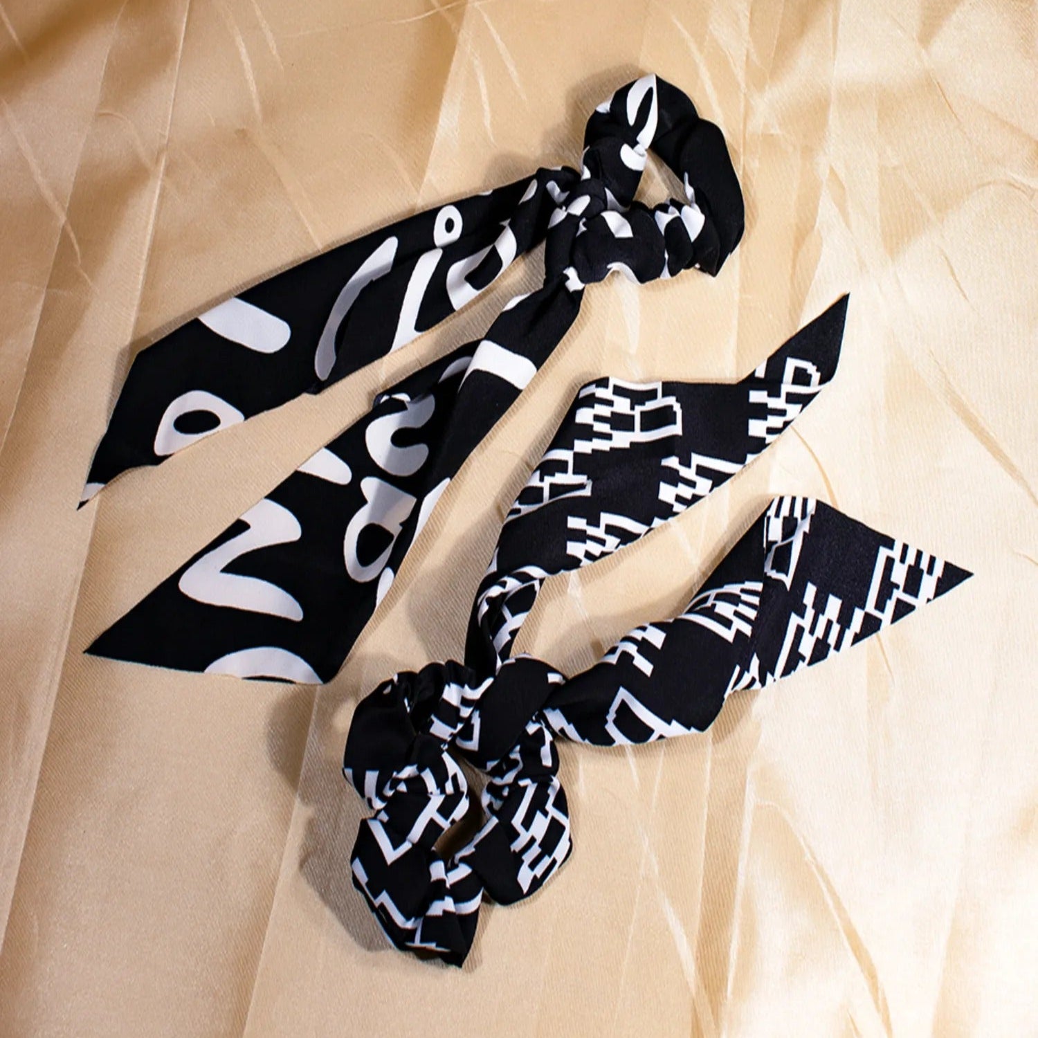 Two black scarf scrunchies with a geometric pattern laid out on a beige background.