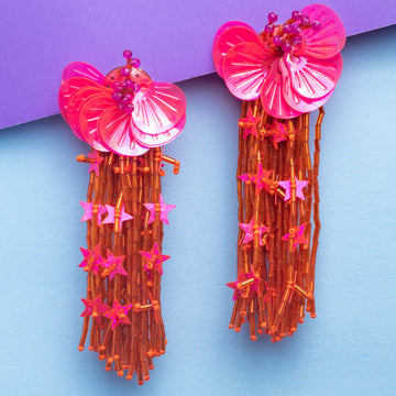 Pair of colorful earrings with pink floral tops and orange tassels on a purple and blue gradient background.