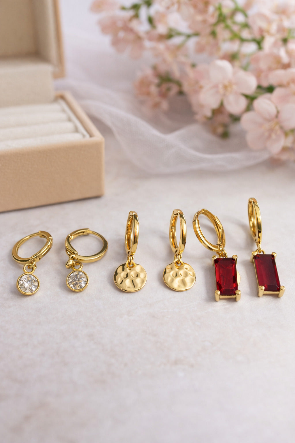 Set of gold hoop earrings with various gemstone drops on a white surface with pink flowers in the background.