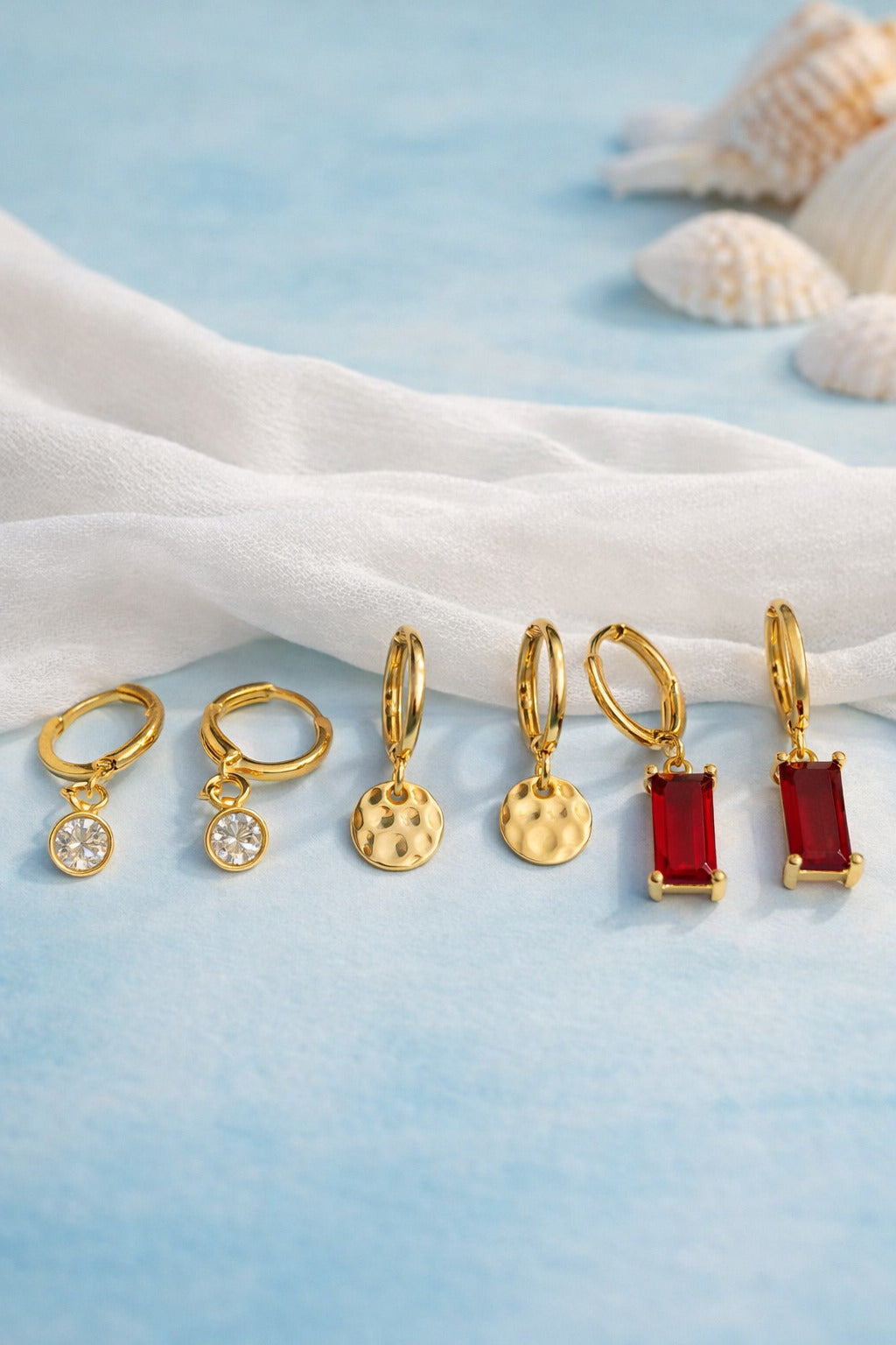 Set of gold and red earrings on a light blue fabric background with seashells.