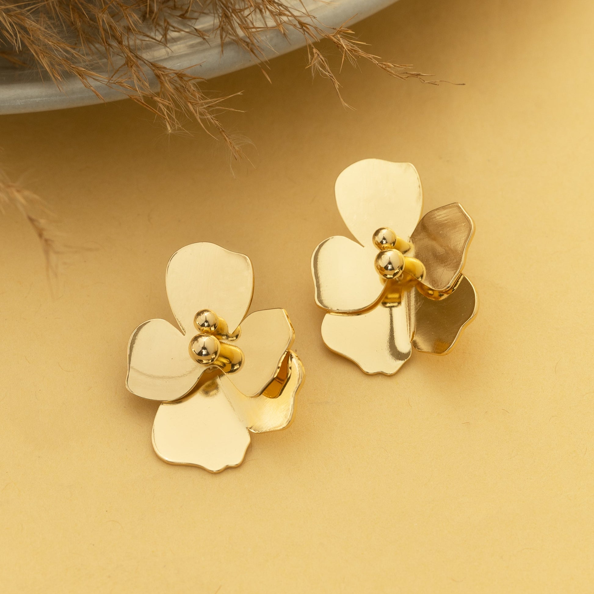 Gold flower-shaped earrings on a beige surface with a plant in the background