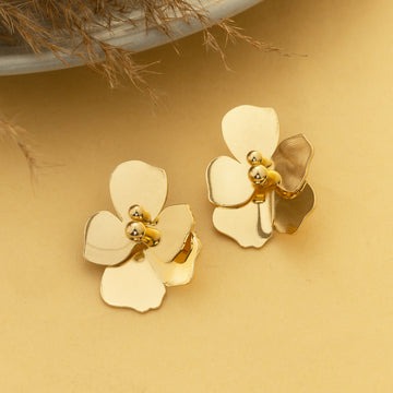 Gold flower-shaped earrings on a beige surface with a plant in the background
