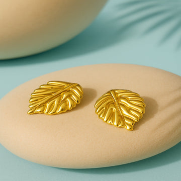 Gold leaf earrings on a beige stone with a light blue background