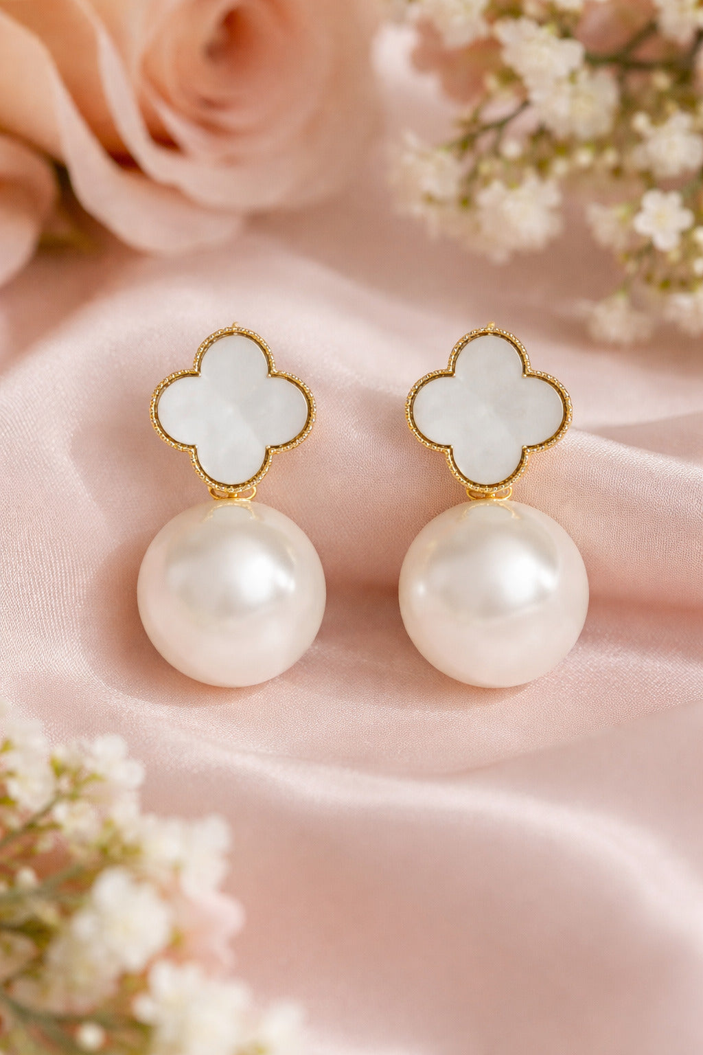 Pearl earrings with gold clover-shaped tops on a soft pink fabric background.