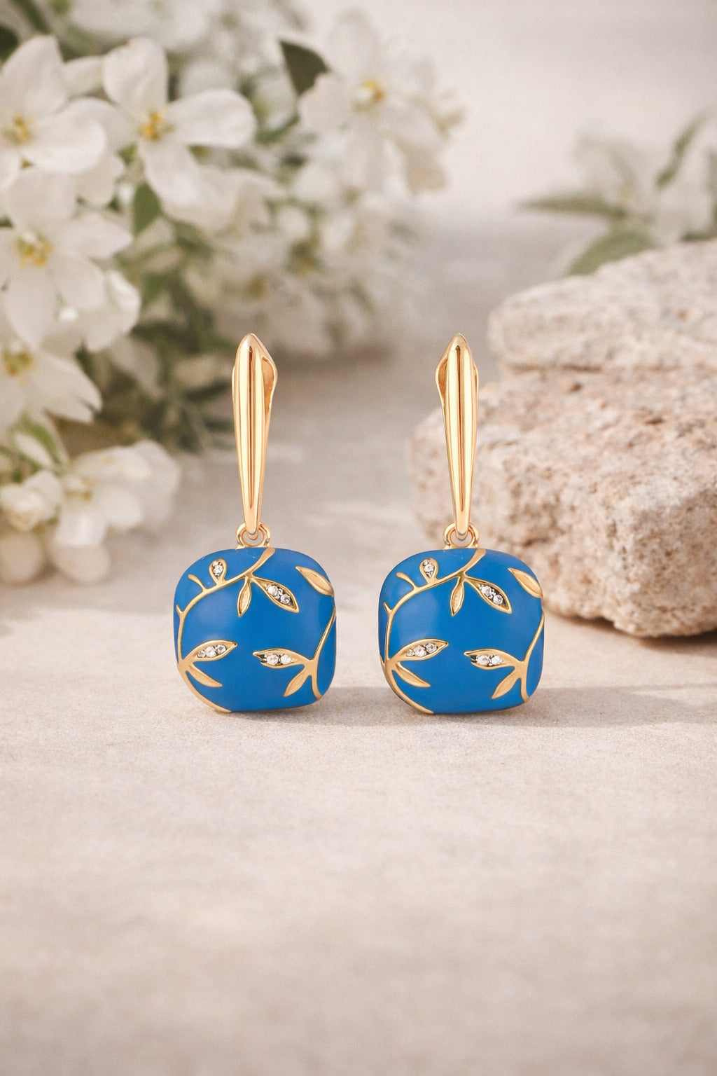 Pair of blue earrings with gold accents on a light surface with white flowers in the background
