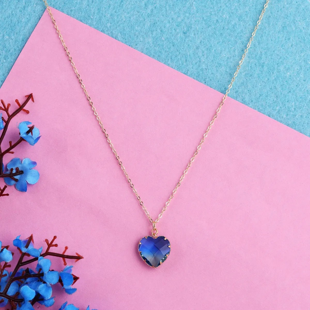 A gold-plated necklace with a blue heart-shaped stone pendant on a chain, displayed against a pink background with blue decorative elements.