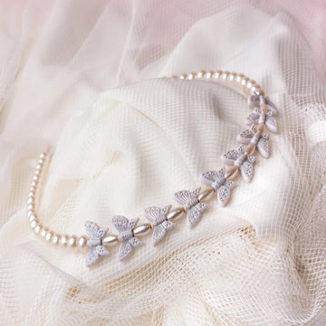 A white hairband with butterfly-shaped designs adorned with imitation pearl beads, displayed on a textured fabric surface.