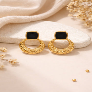 Gold hoop earrings with black square centers on a beige surface with dried flowers.