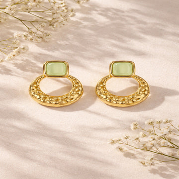 Gold earrings with light green square stones on a beige background with dried flowers.