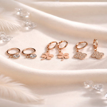 Set of rose gold earrings with leaf and heart designs on a soft fabric background