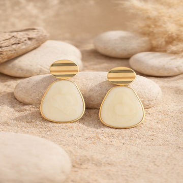 Gold and cream earrings on a sandy surface with stones in the background