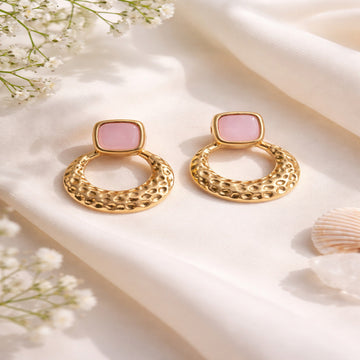 Gold earrings with pink stones on a soft white background with flowers and a shell.