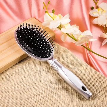 A plastic hair comb with a curved handle and round bristle head, placed on a beige surface with a wooden comb and white flowers in the background.