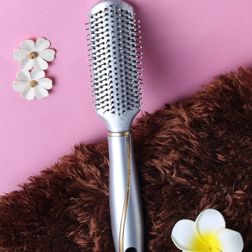 A silver-colored Jewelz trendy hair brush with round bristles, shown alongside a brown furry surface and decorative white flowers on a pink background.