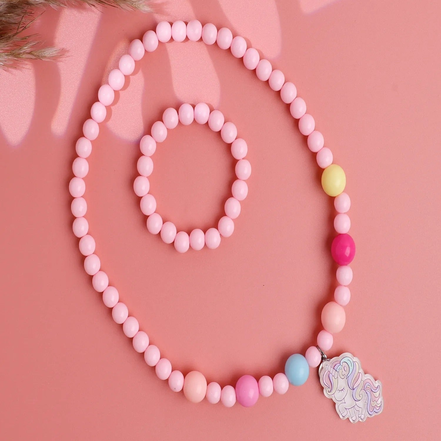 A set of plastic pink kids necklace and bracelet jewelry with a cute charm on the necklace, displayed on a pink background.