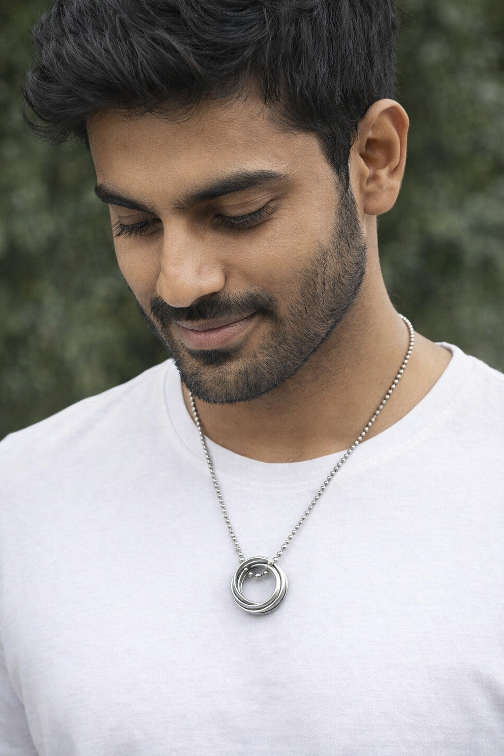 Man wearing a silver necklace with a circular pendant against a blurred green background