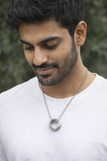Man wearing a silver necklace with a circular pendant against a blurred green background