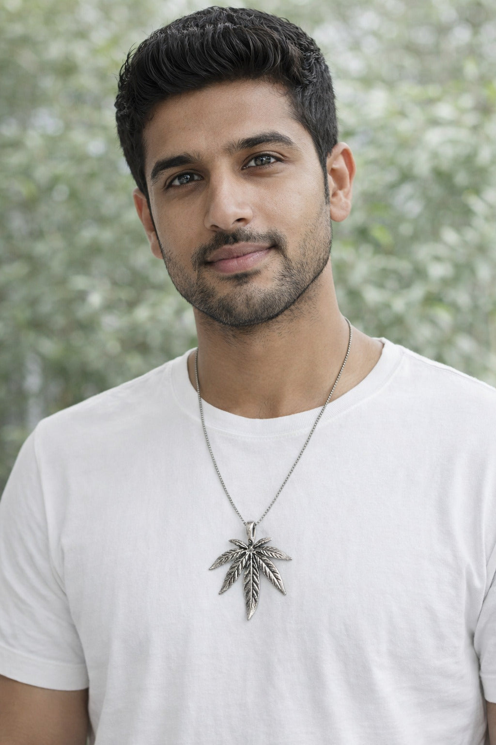 Man wearing a white t-shirt with a necklace featuring a leaf pendant against a blurred green background