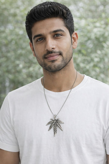 Man wearing a white t-shirt with a necklace featuring a leaf pendant against a blurred green background