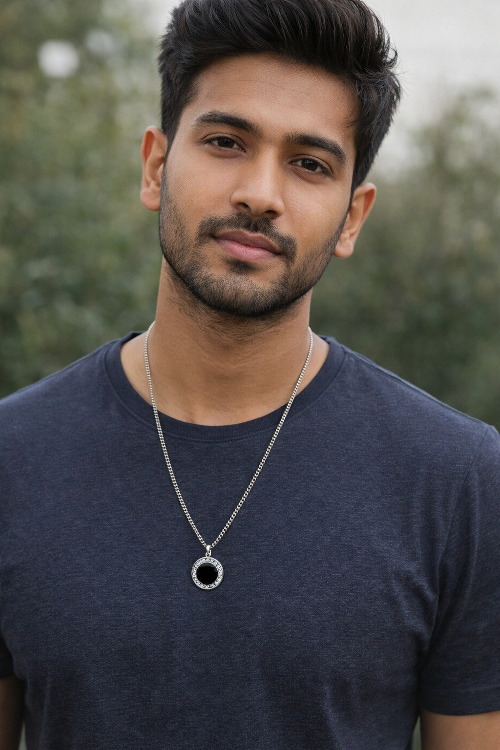 Man wearing a dark blue shirt and a necklace with a circular pendant, standing outdoors.