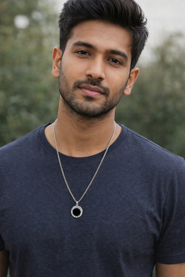 Man wearing a dark blue shirt and a necklace with a circular pendant, standing outdoors.