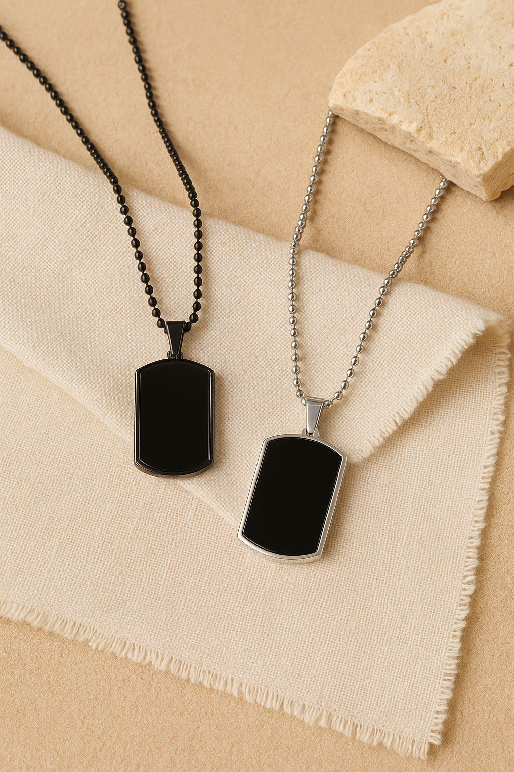 Two black dog tag necklaces on a beige fabric background