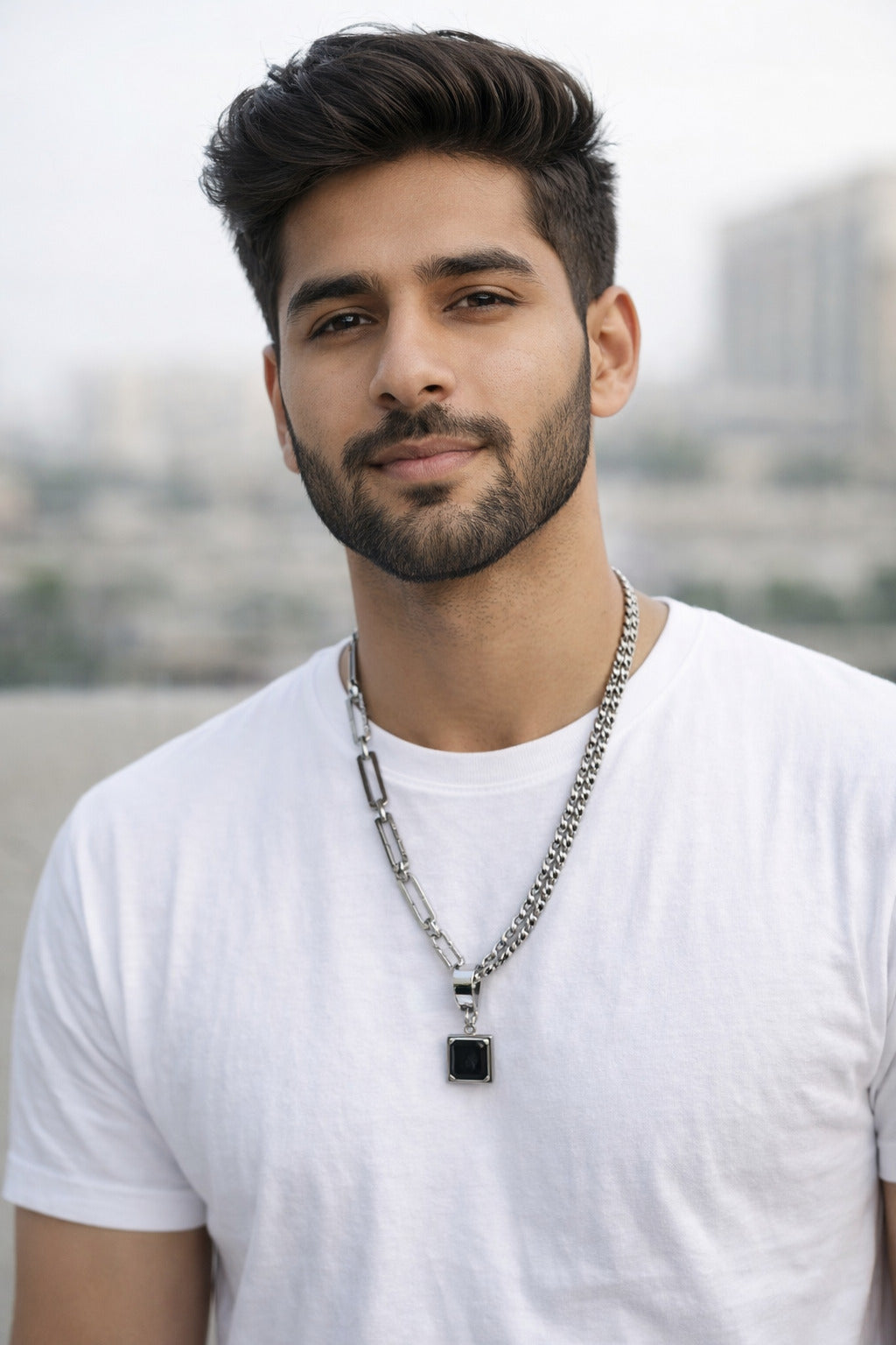 Man wearing a white t-shirt and silver necklace with a black pendant, standing outdoors.