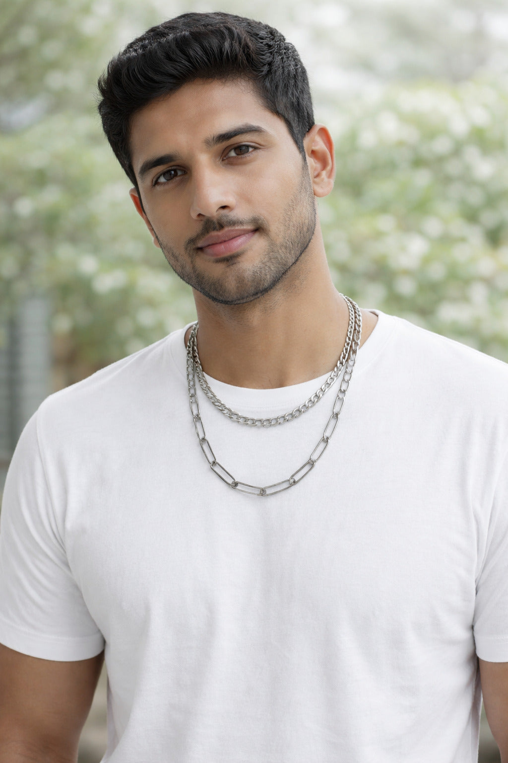 Man wearing a white t-shirt and multiple chain necklaces with a blurred natural background