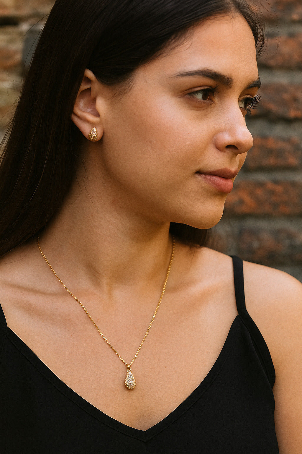 Woman wearing a gold necklace with a teardrop pendant against a brick wall.