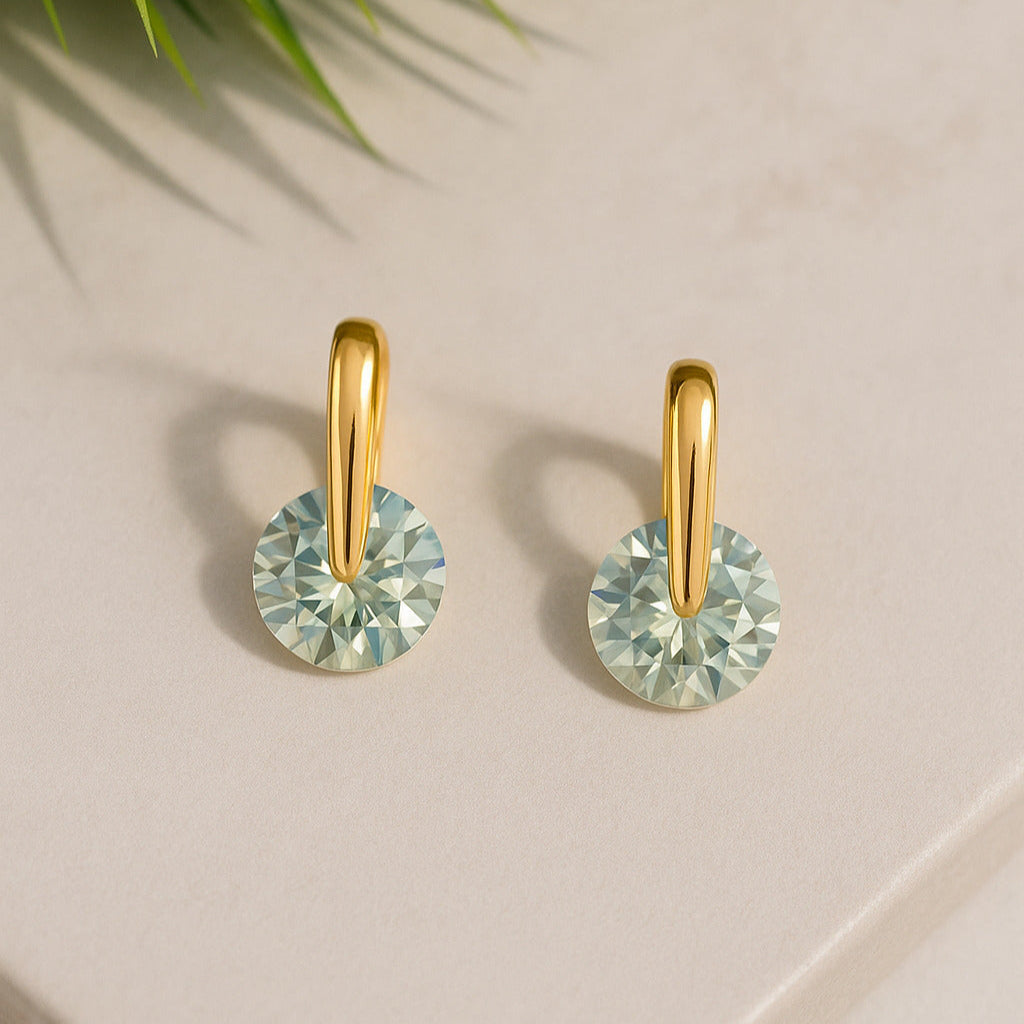 Gold earrings with green gemstones on a light surface with a plant leaf in the corner.