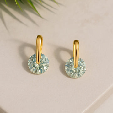 Gold earrings with green gemstones on a light surface with a plant leaf in the corner.
