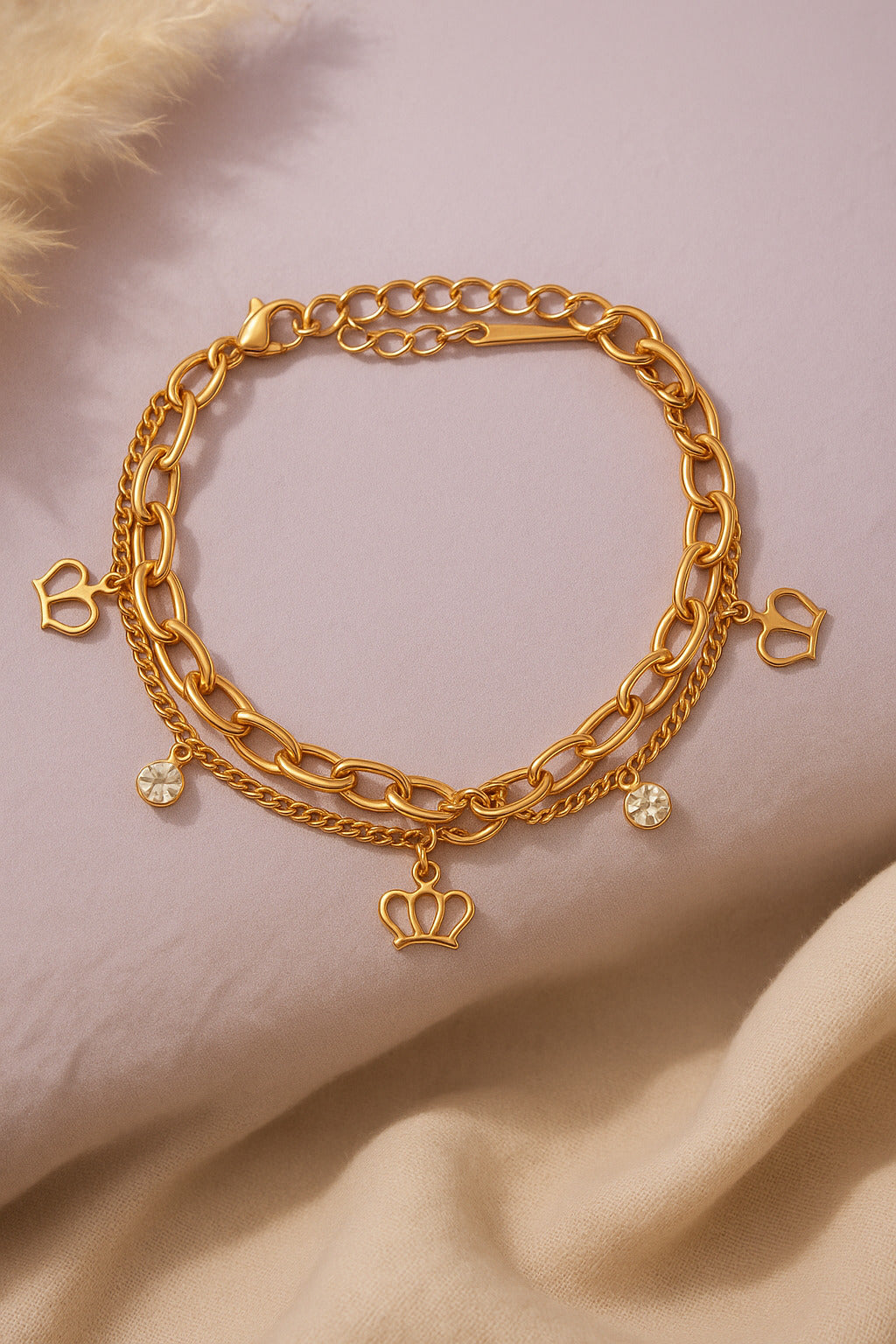 Gold chain bracelet with heart and crown charms on a beige fabric background