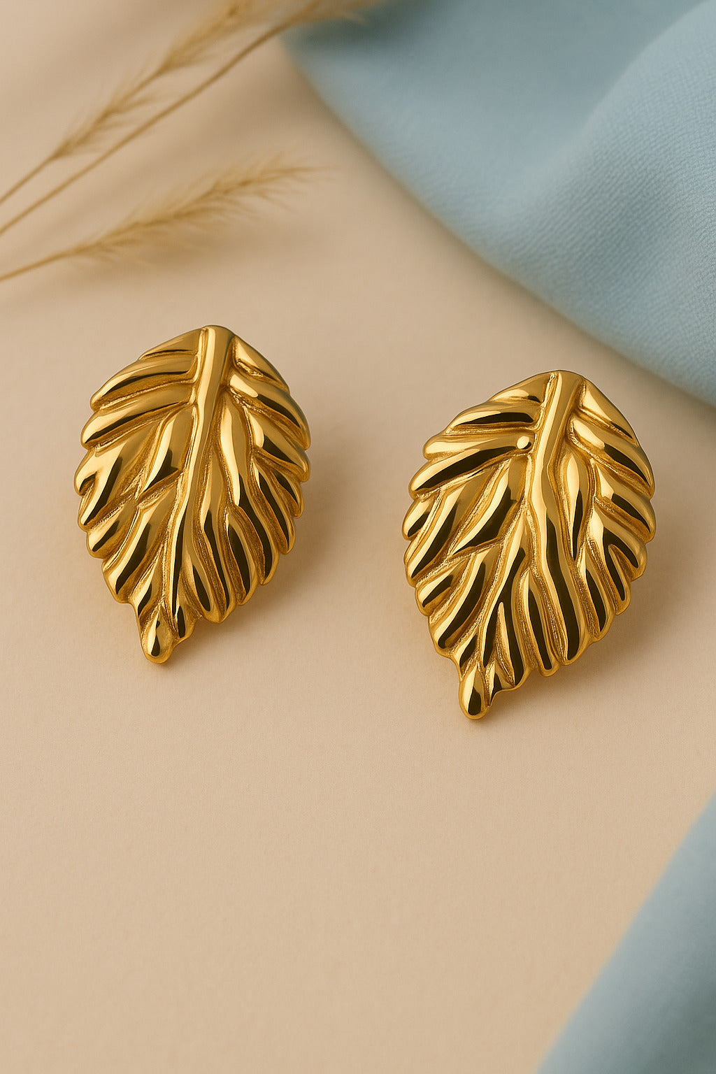 Gold leaf-shaped earrings on a beige surface with a blue fabric background