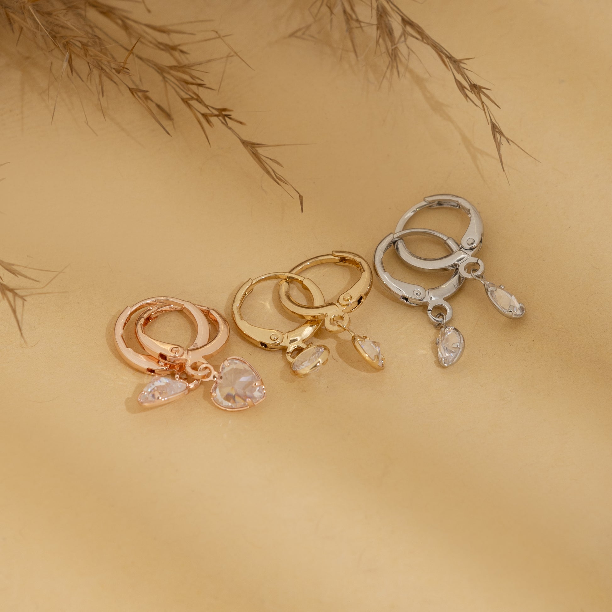 Three gold hoop earrings with heart and teardrop charms on a beige background