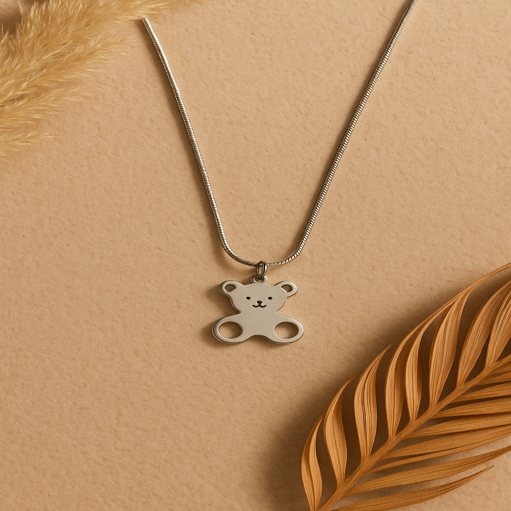 Silver teddy bear pendant necklace on a beige background with dried leaves.