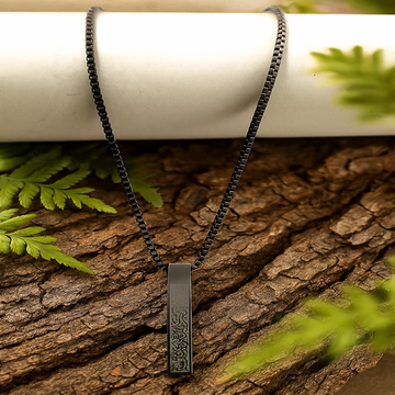 Black chain necklace with a rectangular pendant on a wooden surface with green leaves.