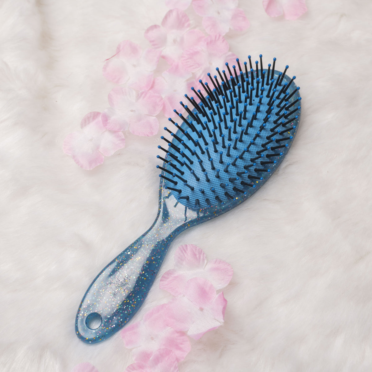 A blue Jewelz trendy hair brush with bristles, placed on a surface with pink petals around it.